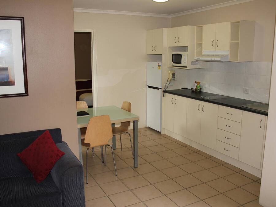 A compact kitchen with white cabinets, a microwave, and a refrigerator. There is a dining table with chairs and a red pillow on a couch in the foreground.