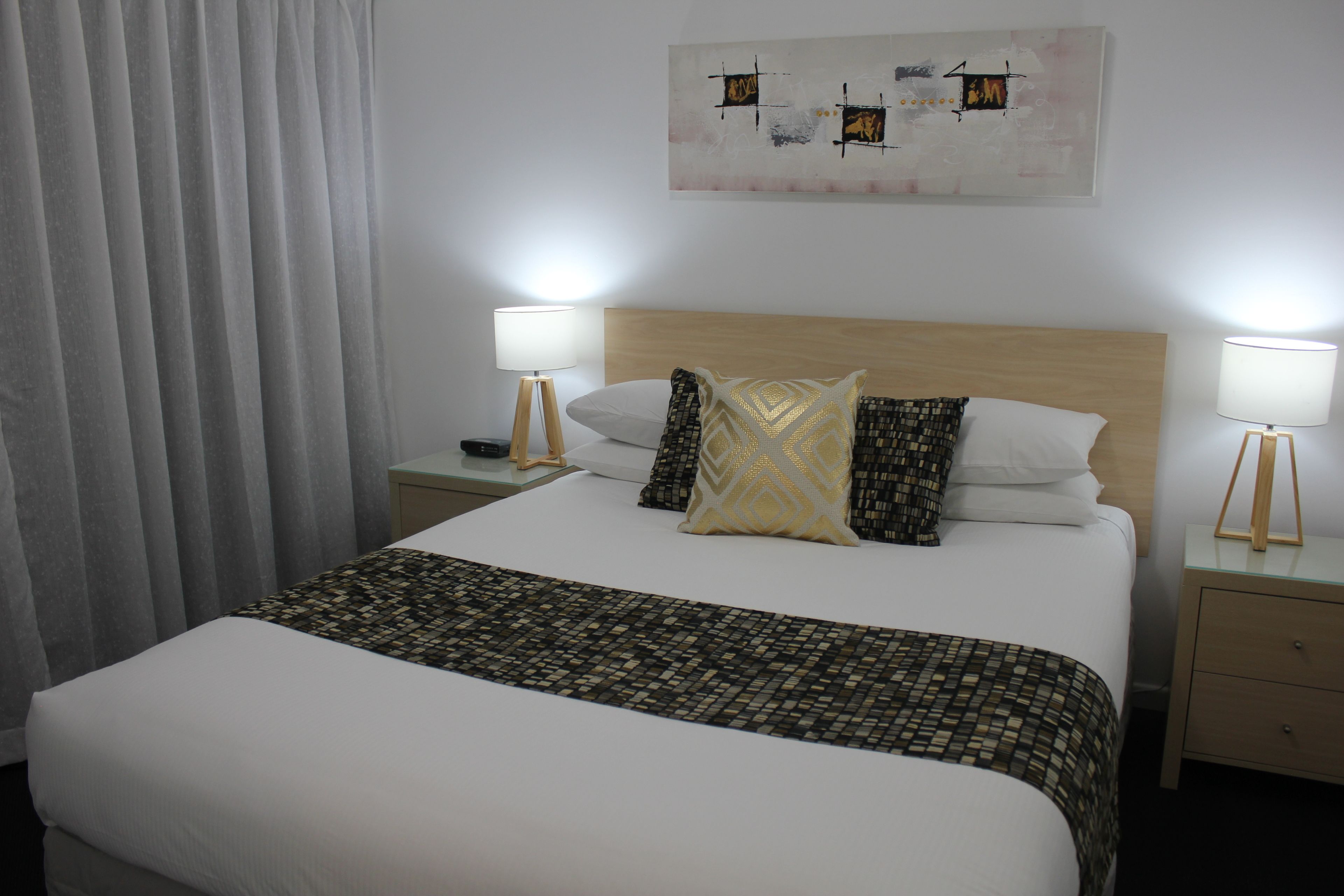 A neatly made bed with a patterned bedspread, two bedside lamps, and a wall-mounted abstract painting.