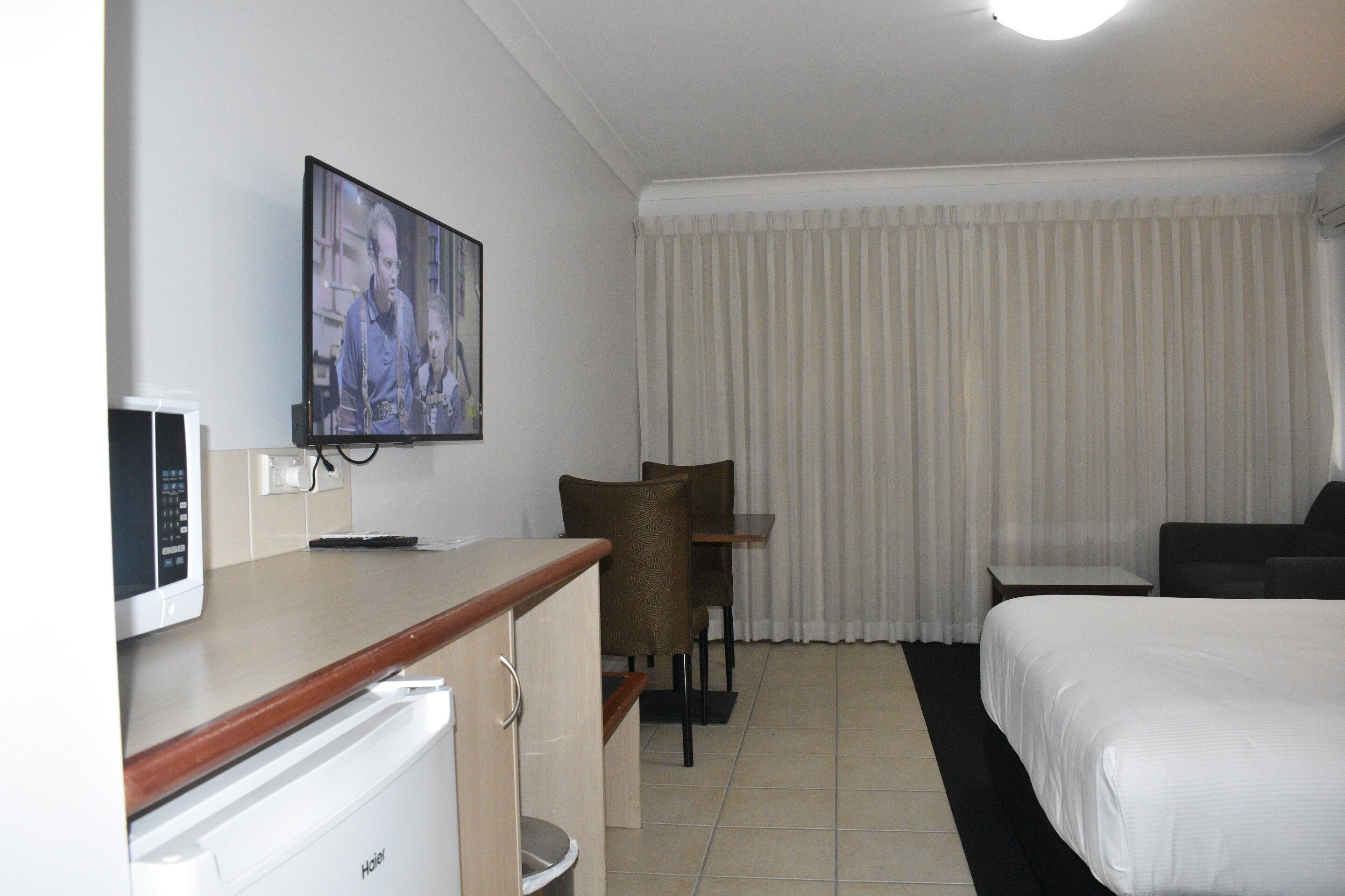 Una habitación de hotel con una cama, un escritorio con microondas, una silla, televisor y ventana con cortinas.