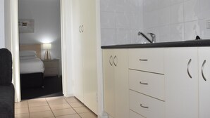 Una habitación de hotel con cama, mesita de noche con lámpara, kitchenette con armarios y baño visible a través de una puerta abierta.