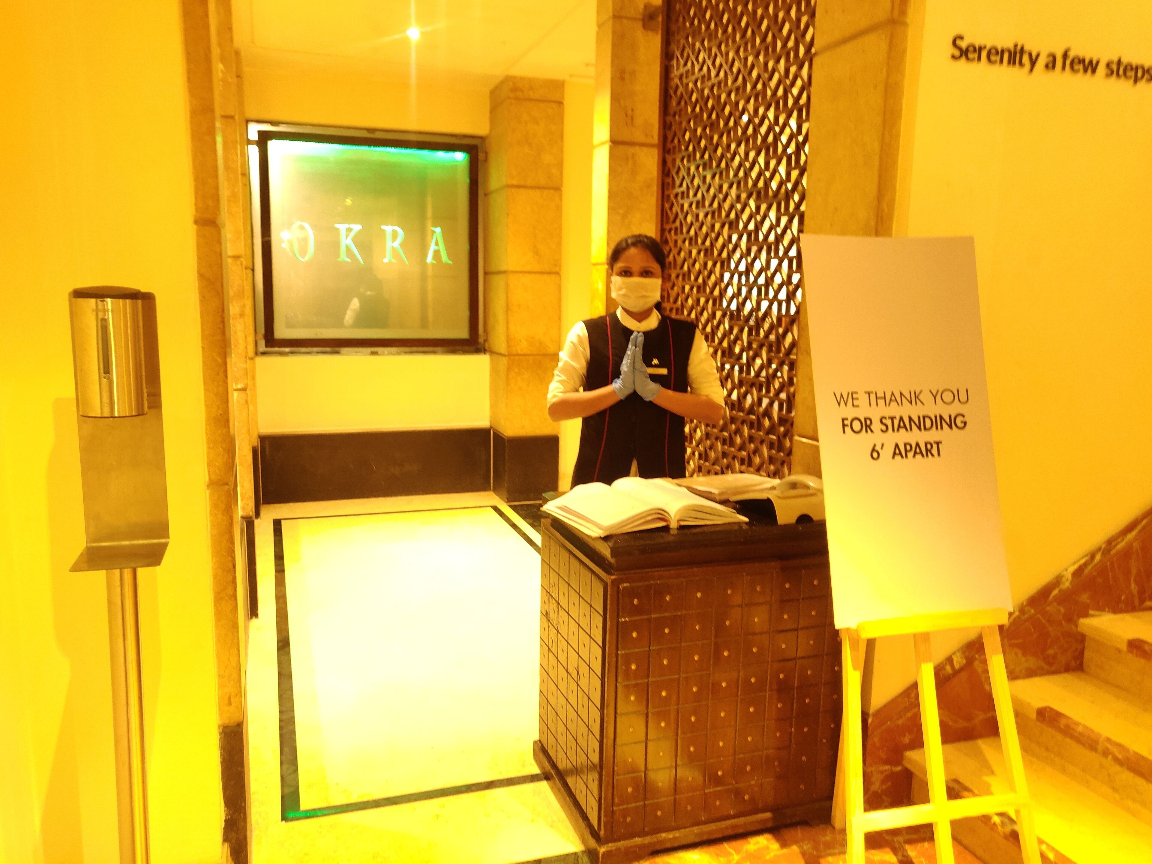 A hotel reception area with a sign reading "Serenity a few steps away" and a message on a stand thanking guests for standing 6 feet apart.