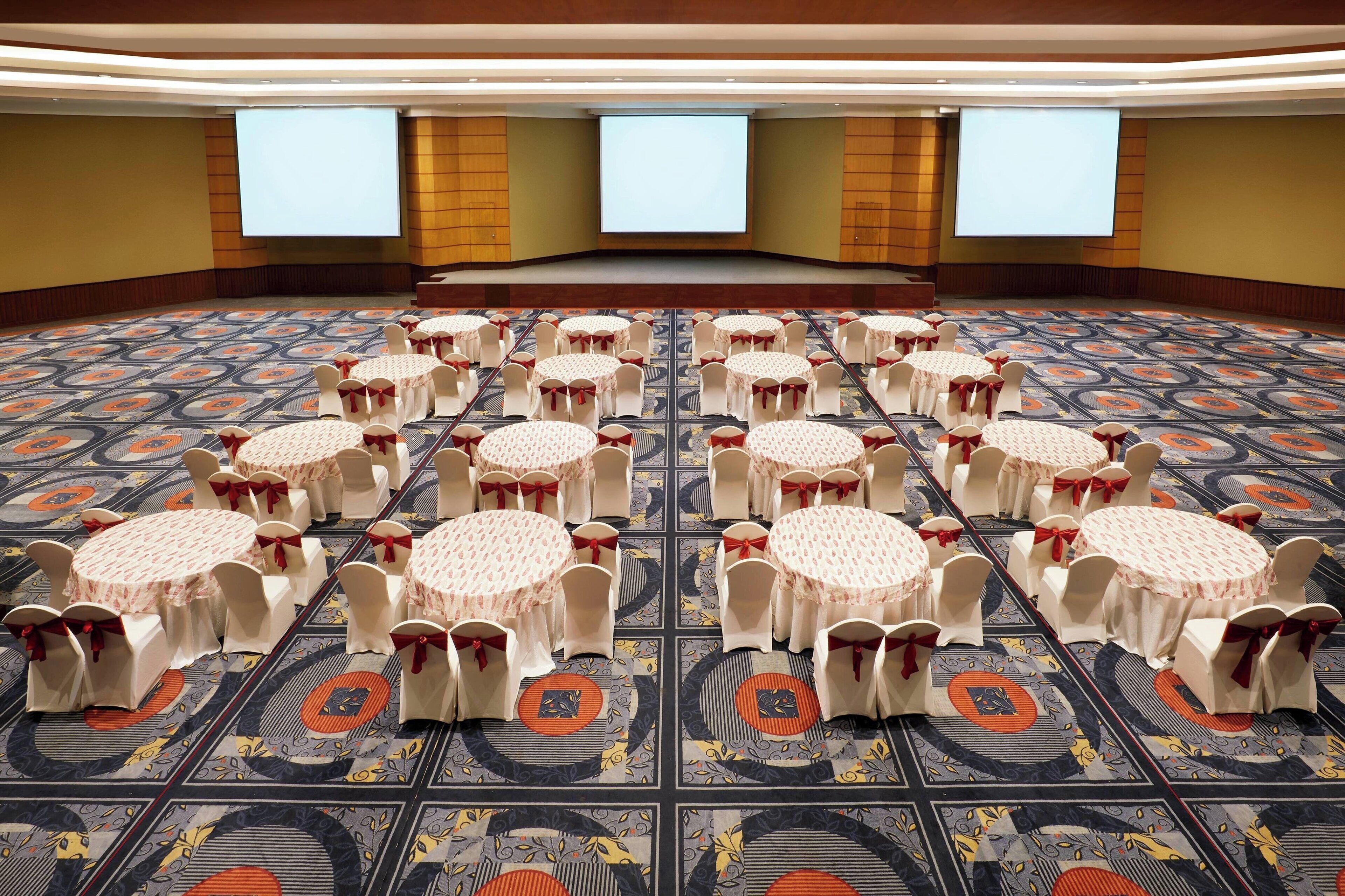 Un salón de conferencias con varias mesas redondas y sillas, cada una con mantel y una cinta roja. La sala tiene una alfombra estampada y grandes pantallas de proyección en la pared de fondo.
