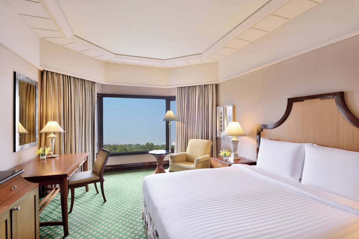 Een hotelkamer met een groot bed, een bureau met stoel, een klein tafeltje en een stoel. Er is een raam met gordijnen, een lamp op het bureau en een lamp op het tafeltje.