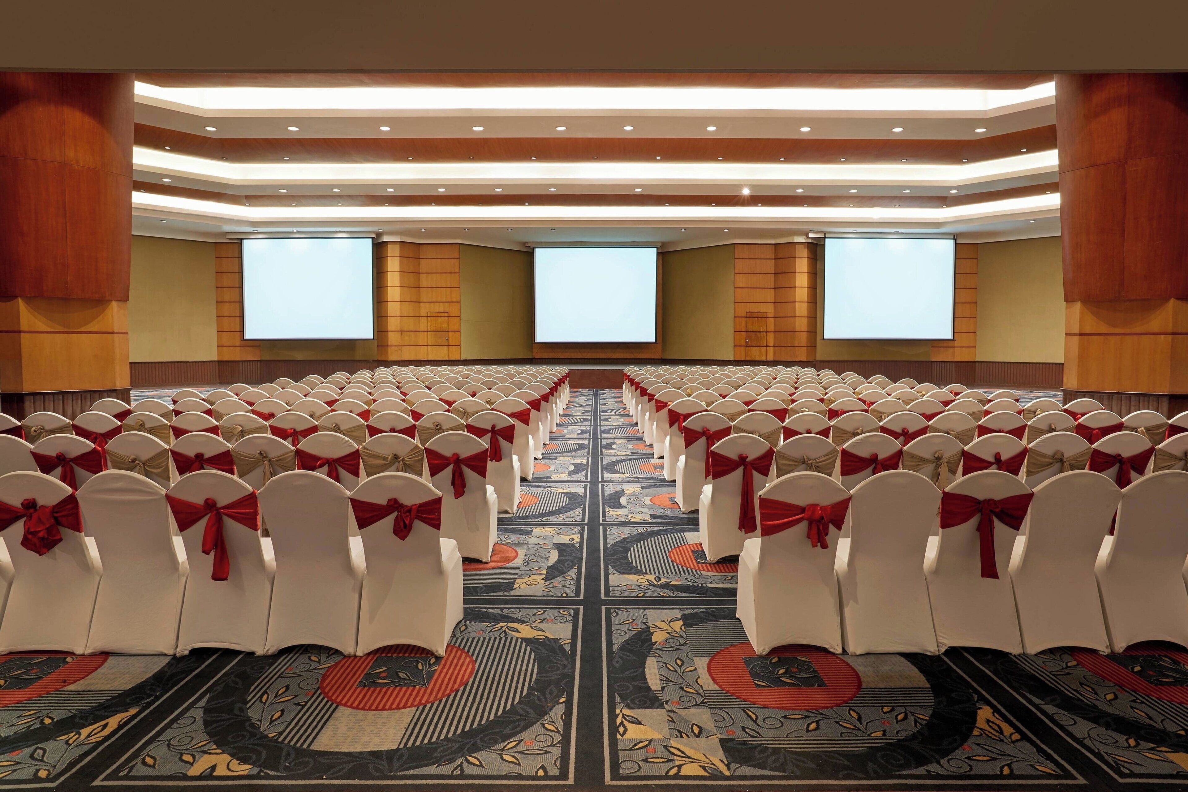 Un salón de conferencias con filas de sillas blancas, cada una decorada con un lazo rojo, y una pantalla grande al frente.