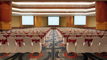 Un salón de conferencias con filas de sillas blancas, cada una decorada con un lazo rojo, y una pantalla grande al frente.