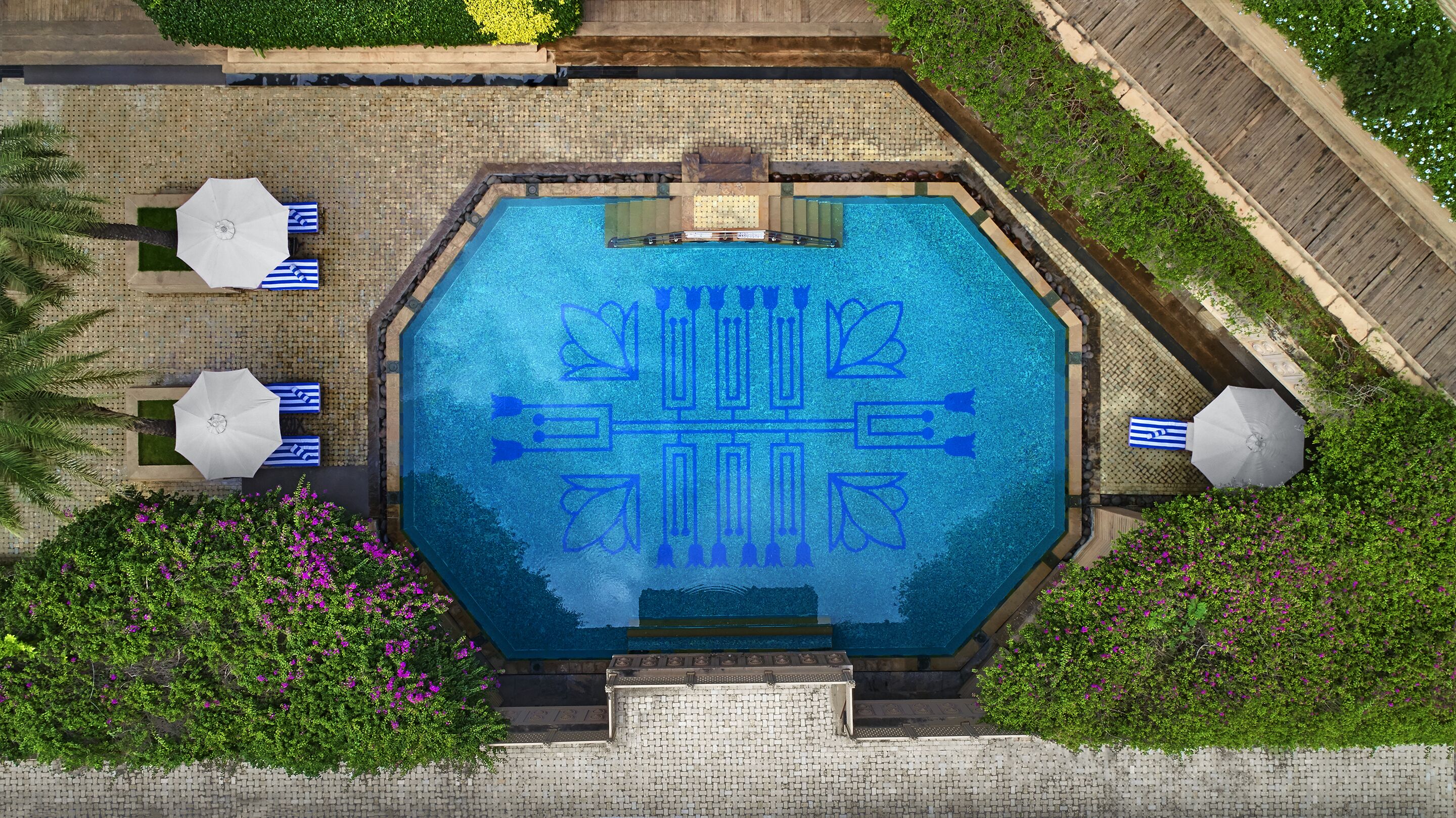 Ein achteckiger Pool mit geometrischem Muster auf dem Boden, umgeben von einer gefliesten Terrasse, Palmen und violetten Blumen.