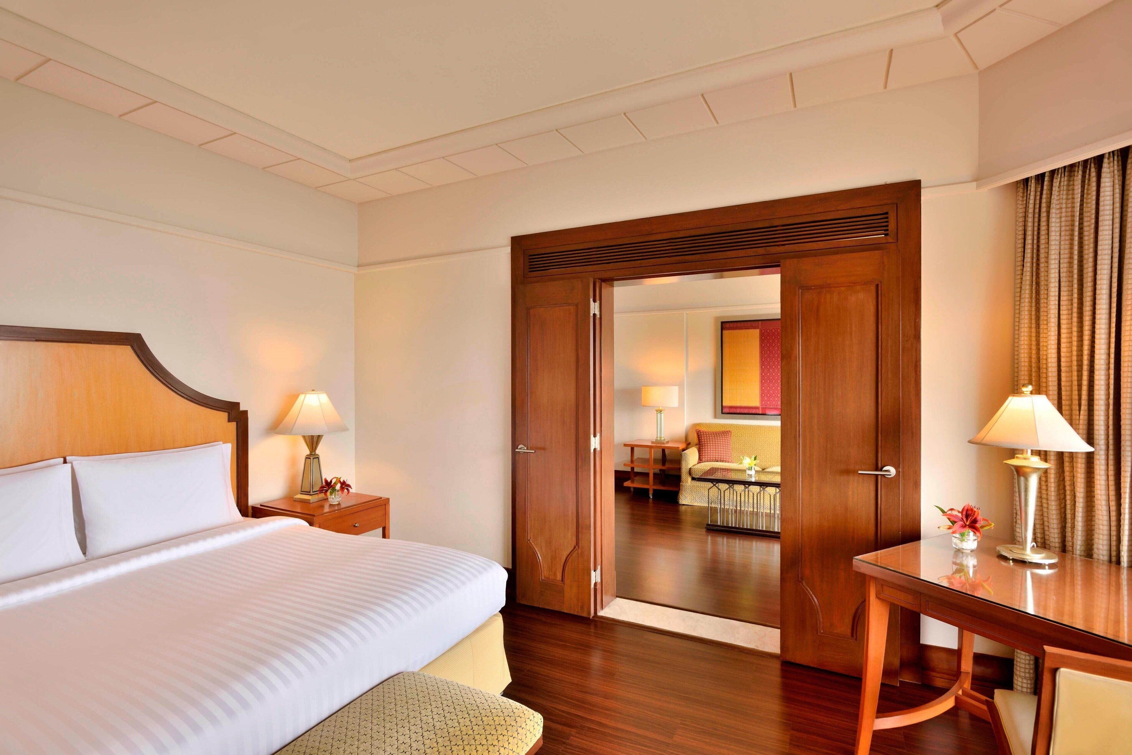 Habitación de hotel con una cama grande, un escritorio de madera y una puerta que da a un área de estar con una mesa y sillas.