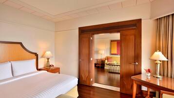 A hotel room with a large bed, a wooden desk, and a door leading to a sitting area with a table and chairs.