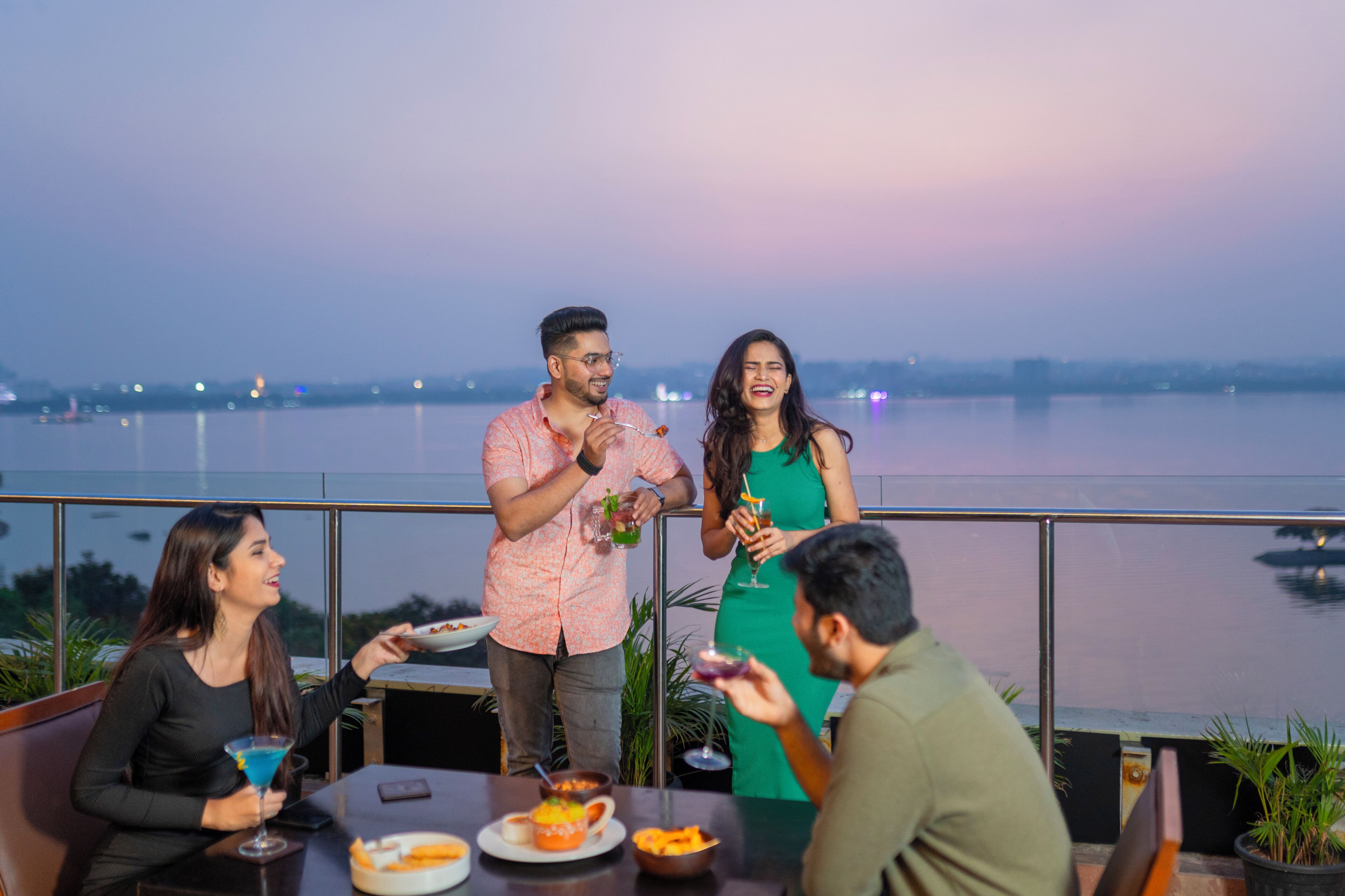 A group of people dining at a table with a view of a body of water and city lights in the background.