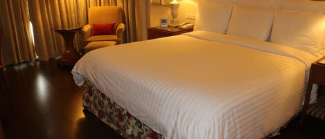 Una habitación de hotel con una cama grande, una silla, una mesita de noche con lámpara y cortinas.