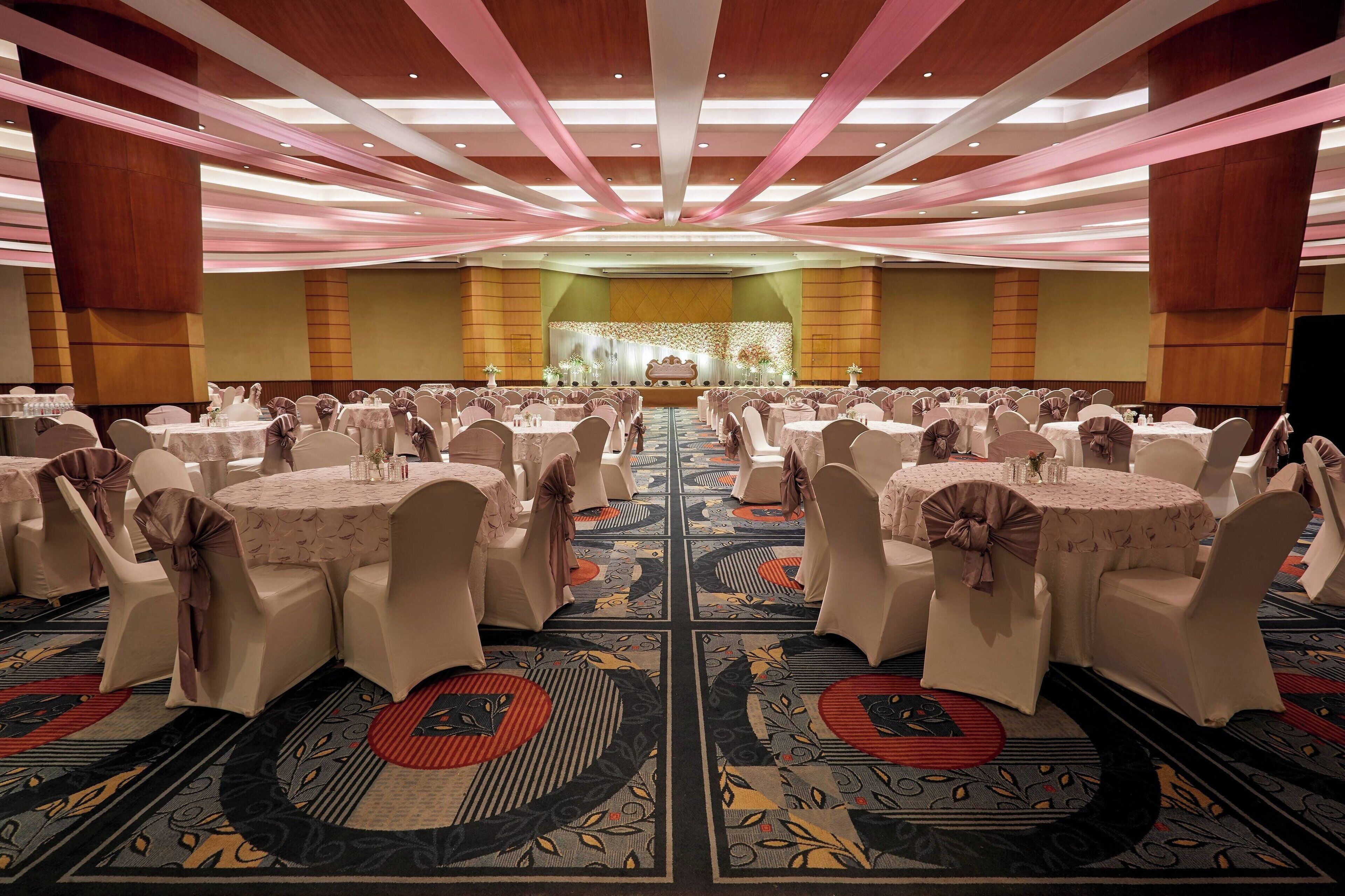 Uma sala de banquetes com mesas redondas, cadeiras brancas e um palco na frente.