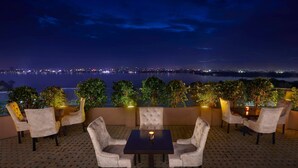 An outdoor seating area with a table and chairs, surrounded by potted plants, overlooking a body of water with city lights in the distance. - Hyderabad Marriott Hotel & Convention Centre (Hyderabad)