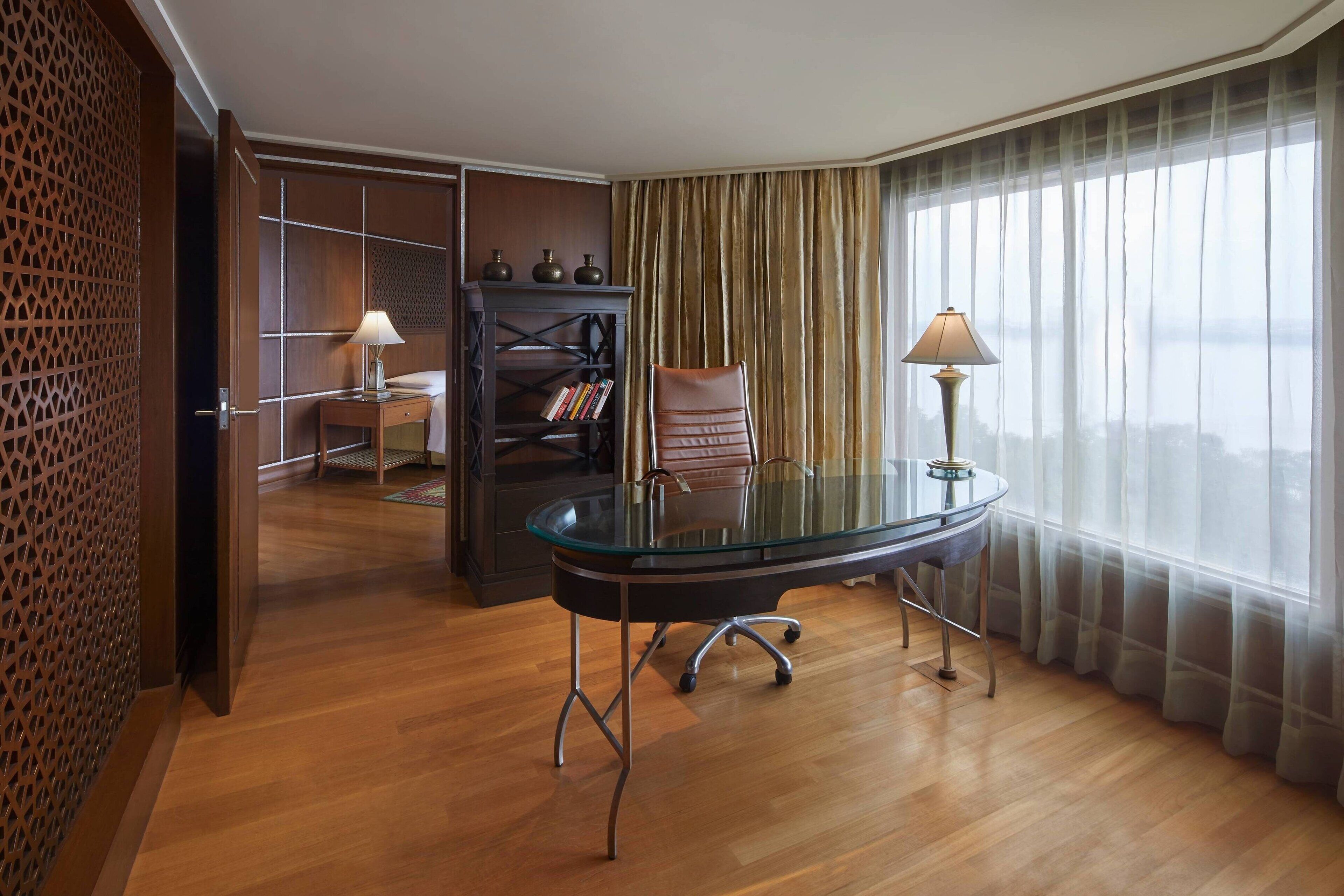 Habitación de hotel con escritorio de vidrio, una silla de cuero, estantería y un ventanal con cortinas traslúcidas.