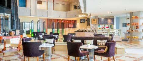 Un vestíbulo amplio con techos altos, una zona de estar central con mesas redondas y sillas moradas, una barra de café y una exhibición de aperitivos y bebidas.