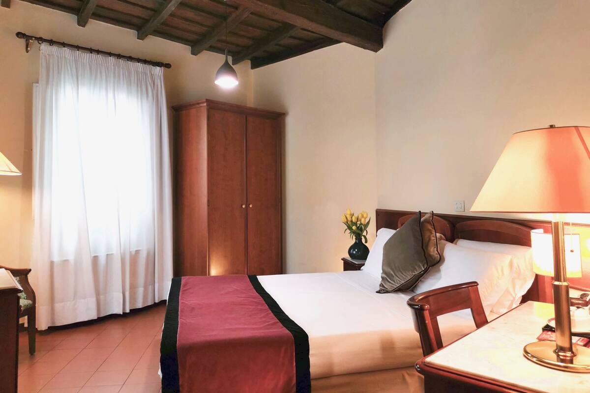 A hotel room with a bed, a wooden wardrobe, a desk with a lamp, and a window with curtains.