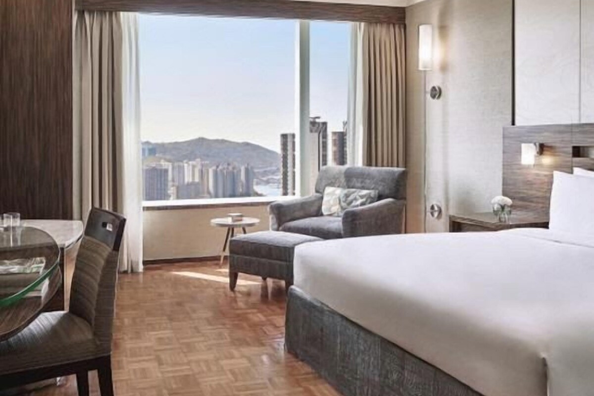 A hotel room with a large bed, a dining table with chairs, a small round table, and a view of a cityscape through a large window.