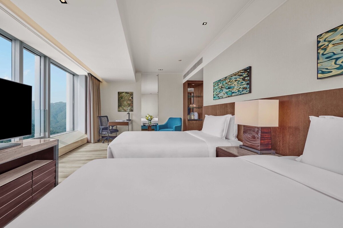 A hotel room with two beds, a television, a desk, and a large window offering a view.
