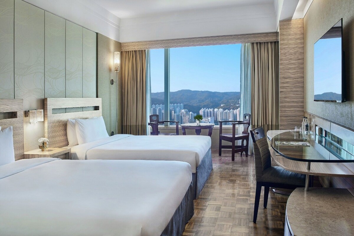 A hotel room with two beds, a dining area, and a large window with a city view.
