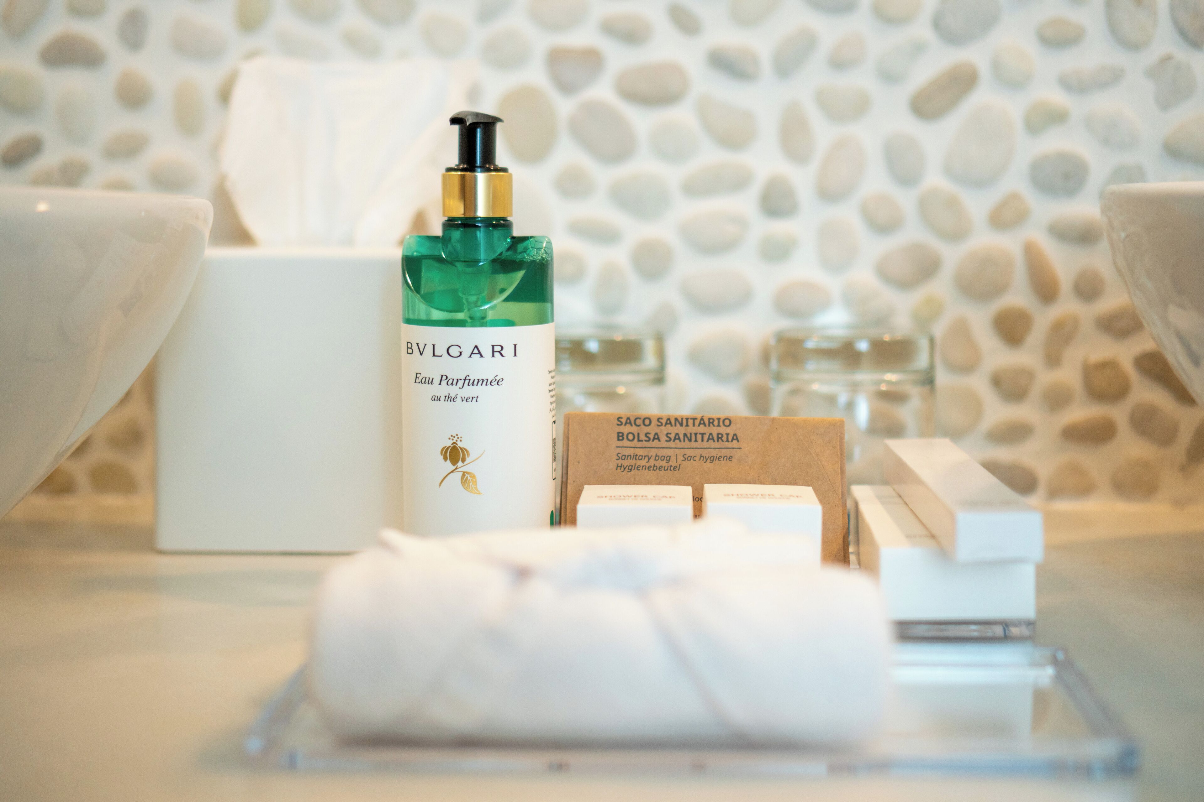 A bathroom with a bottle of BVLGARI Eau Parfumée, a white towel, and a wooden box labeled Saco Sanitário.