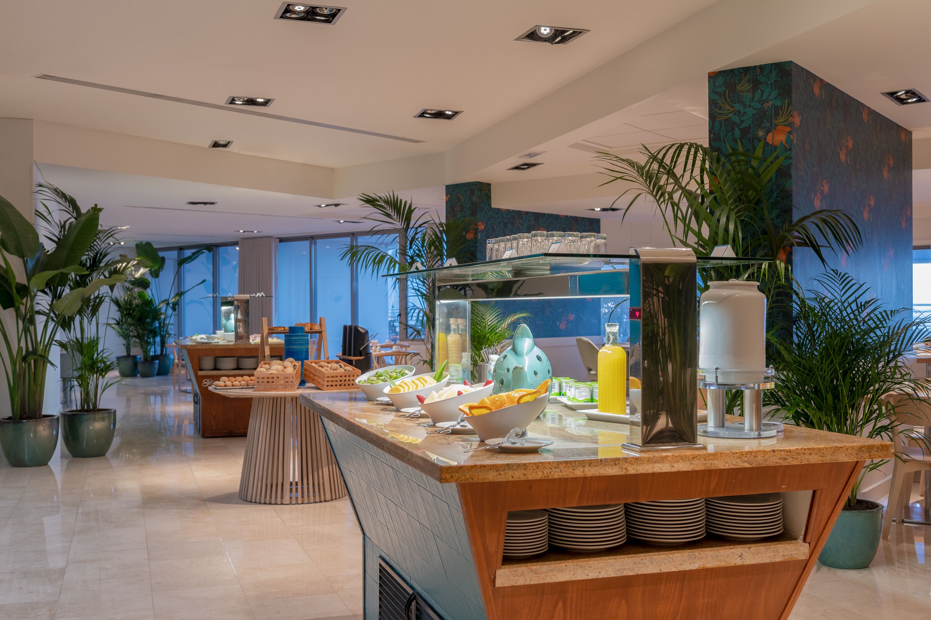 A modern hotel lobby with a breakfast bar, potted plants, and a large window offering a view of the city.