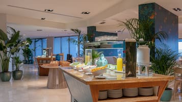 A modern hotel lobby with a breakfast bar, potted plants, and a large window offering a view of the city.