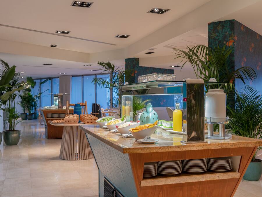 A modern hotel lobby with a breakfast bar, potted plants, and a large window offering a view of the city.