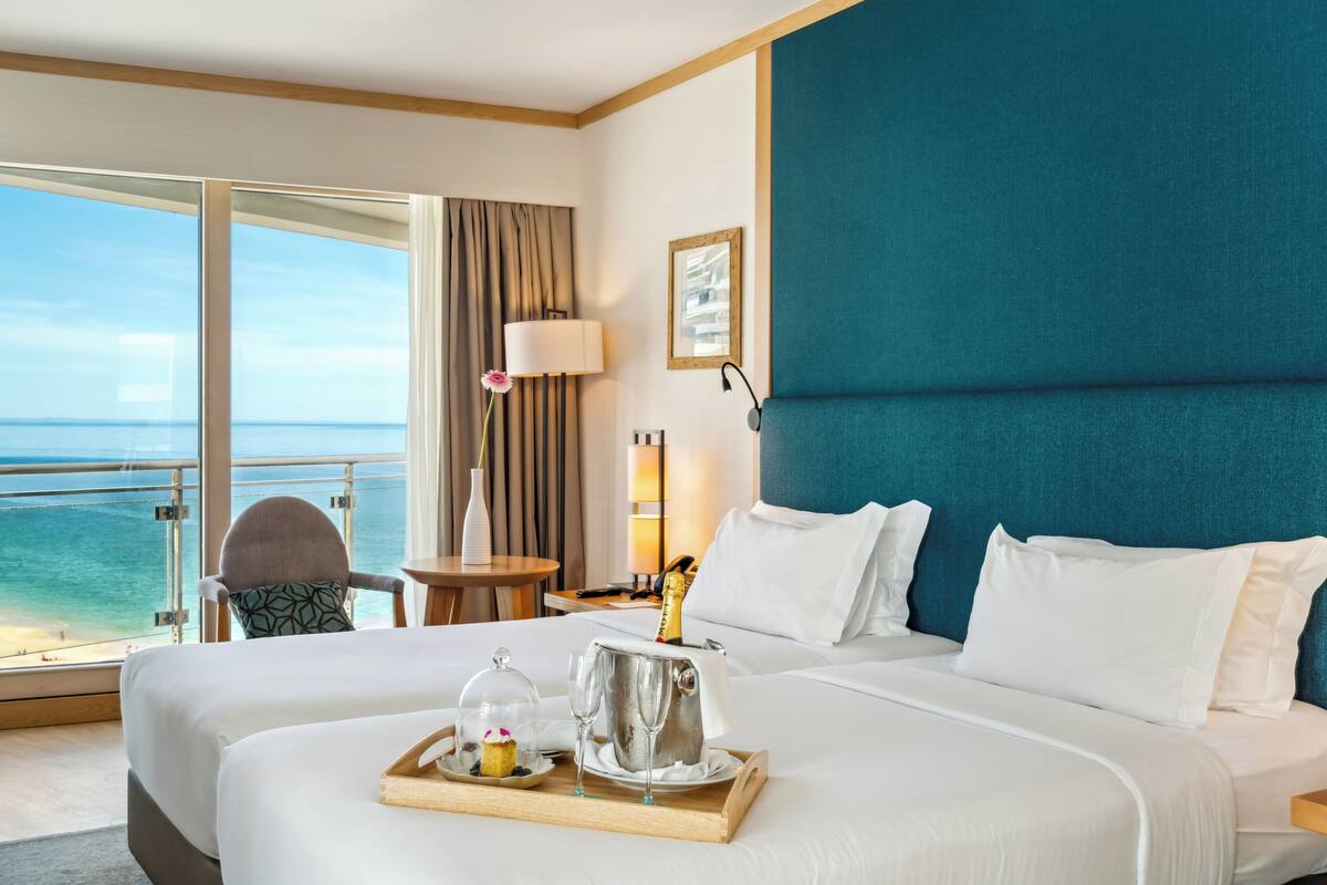Ein Hotelzimmer mit einem großen Bett, Meerblick, einem Balkon mit einem Stuhl und einem Tablett mit Kuchen und Champagnergläsern.
