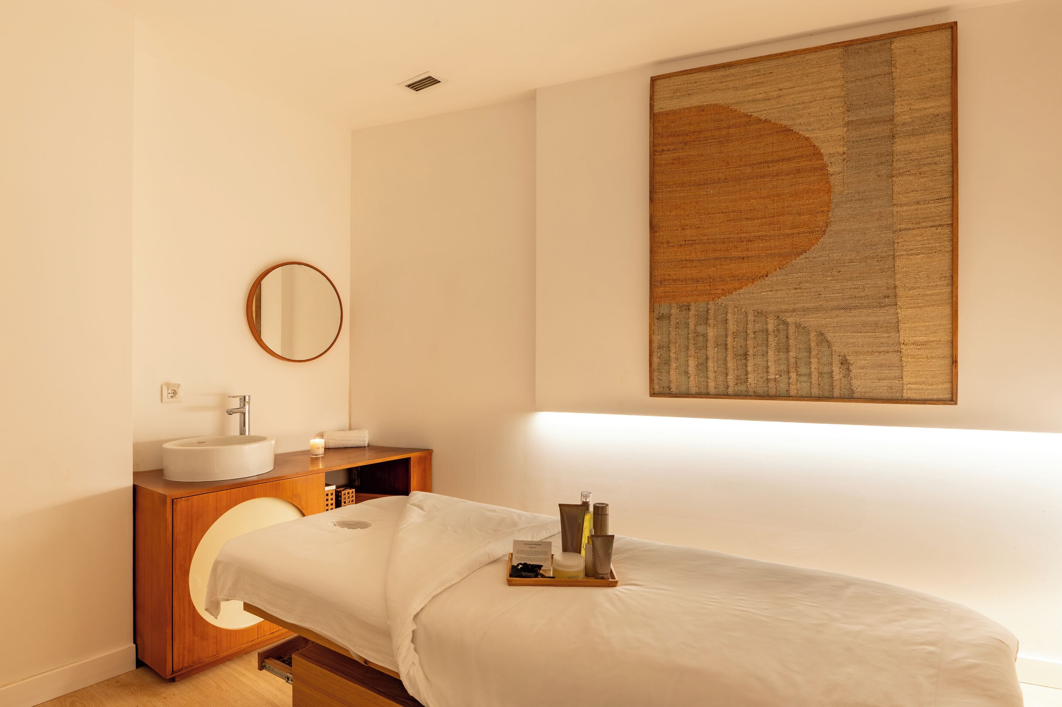 A modern hotel room with a wooden vanity, a round mirror, a bed with a tray of toiletries, and a large abstract painting on the wall.