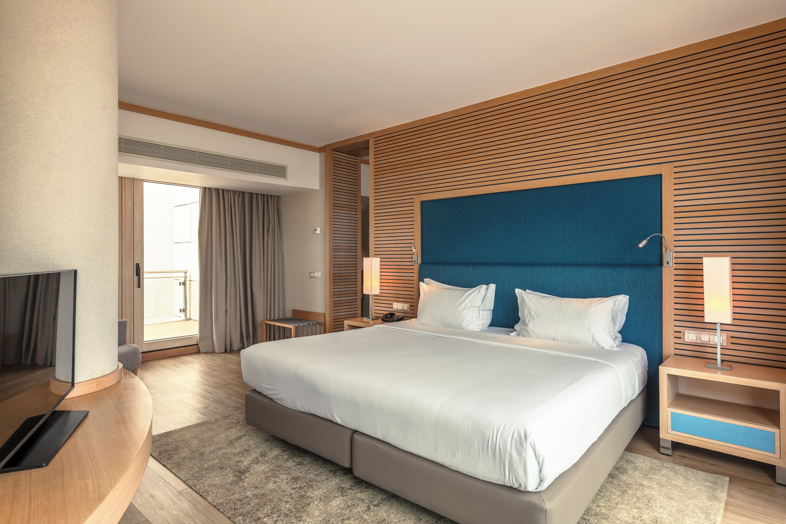 A modern hotel room with a large bed, a TV, a desk, and a balcony view.