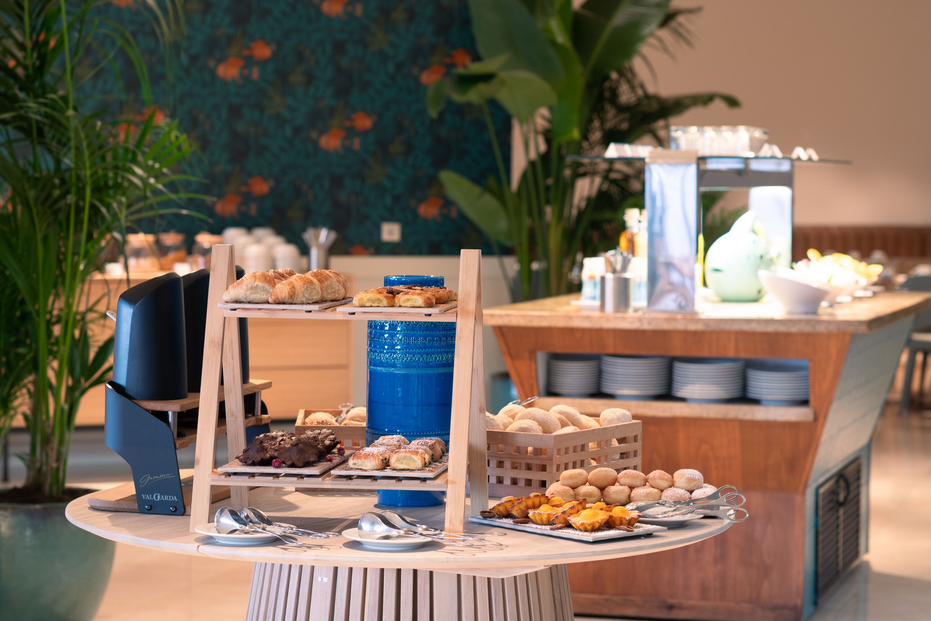 A buffet with various pastries and desserts, a blue water dispenser, and a wooden stand with plates and cutlery.