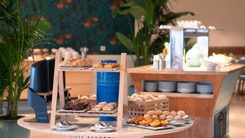 A buffet with various pastries and desserts, a blue water dispenser, and a wooden stand with plates and cutlery.