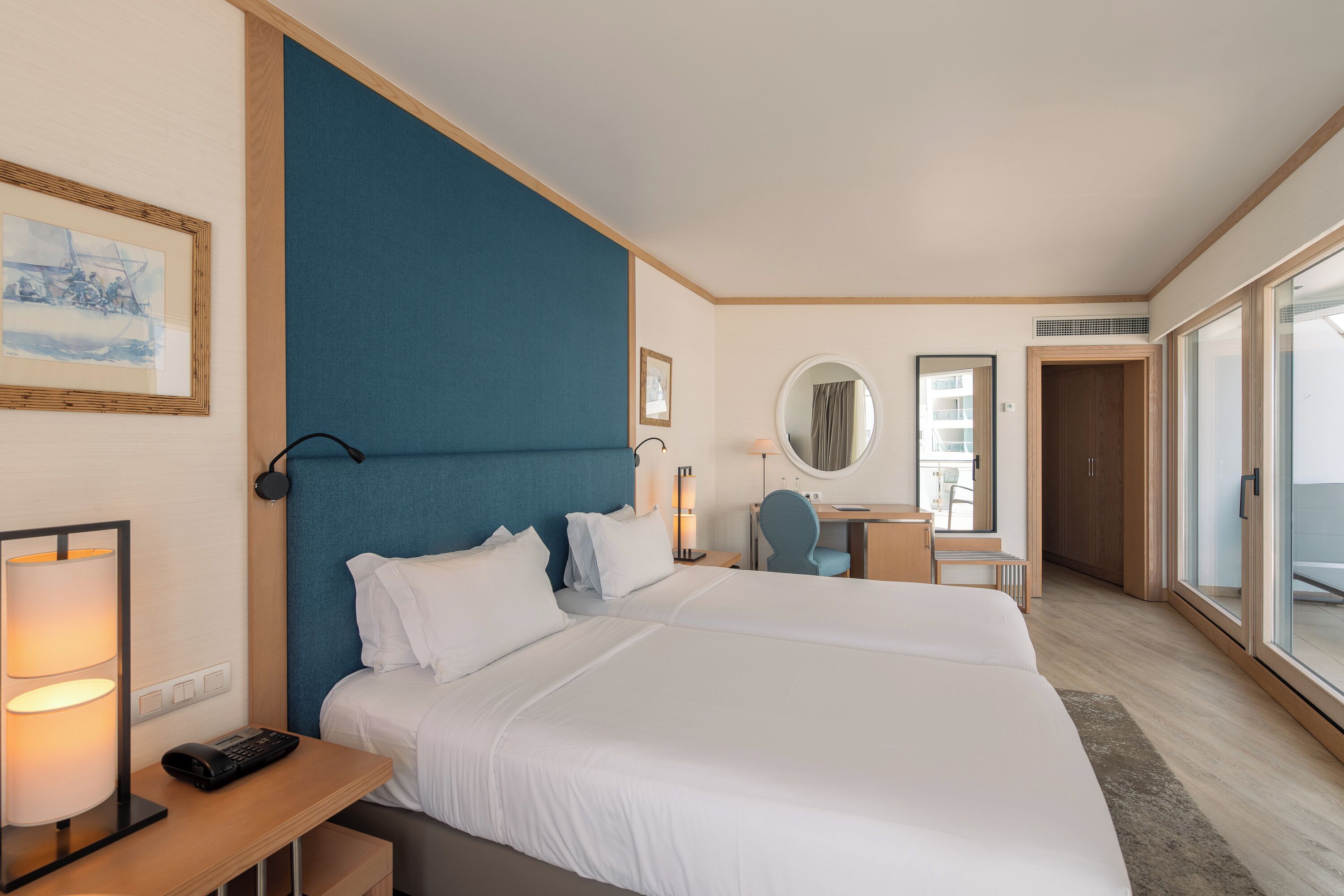 A hotel room with a large bed, a desk with a lamp, a chair, a mirror, and a view of the outside.