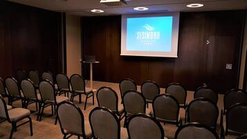 Un salón de conferencias con filas de sillas mirando hacia una pantalla de proyección que muestra
