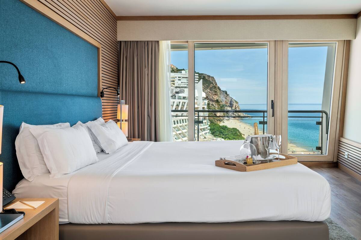 Ein großes Bett mit weißen Leinentüchern, ein Nachttisch mit einer Lampe und ein Tablett mit einer Flasche und Gläsern. Ein großes Fenster bietet Blick auf einen Strand und Gebäude.
