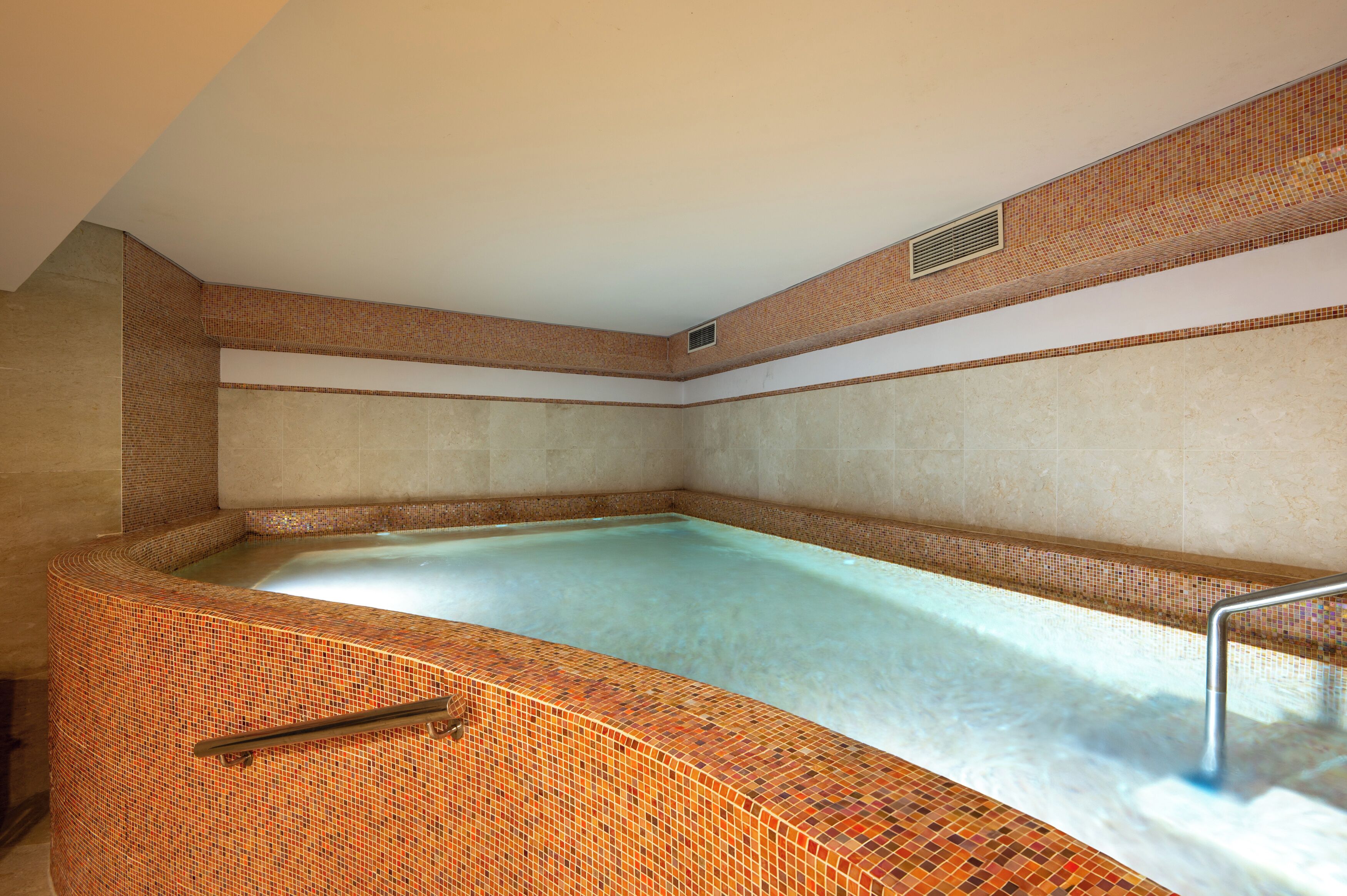 A spa room with a tiled hot tub, a handrail, and a ventilation vent on the ceiling.