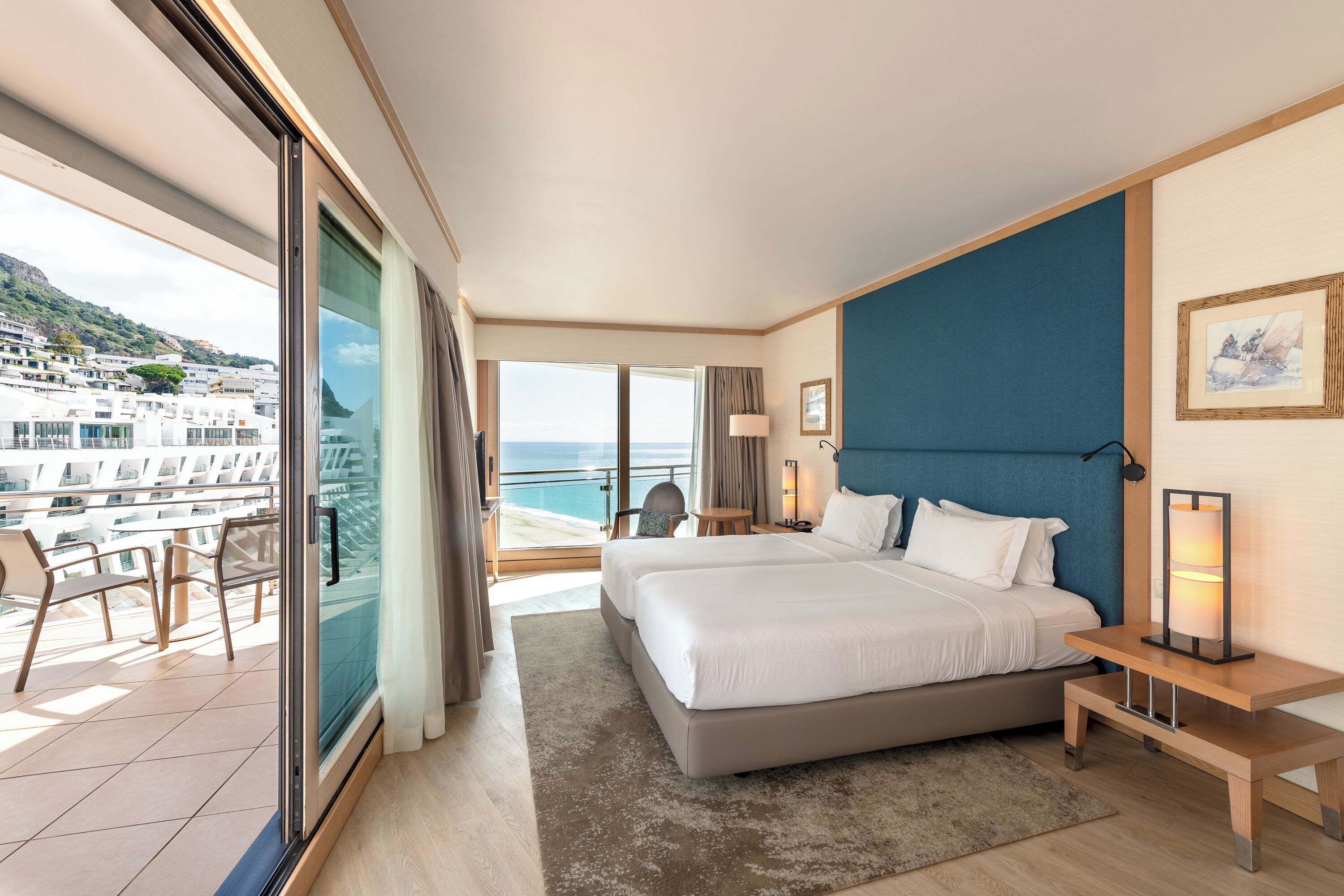 Une chambre d’hôtel avec un grand lit, un balcon avec des chaises et une vue sur la mer et les bâtiments.