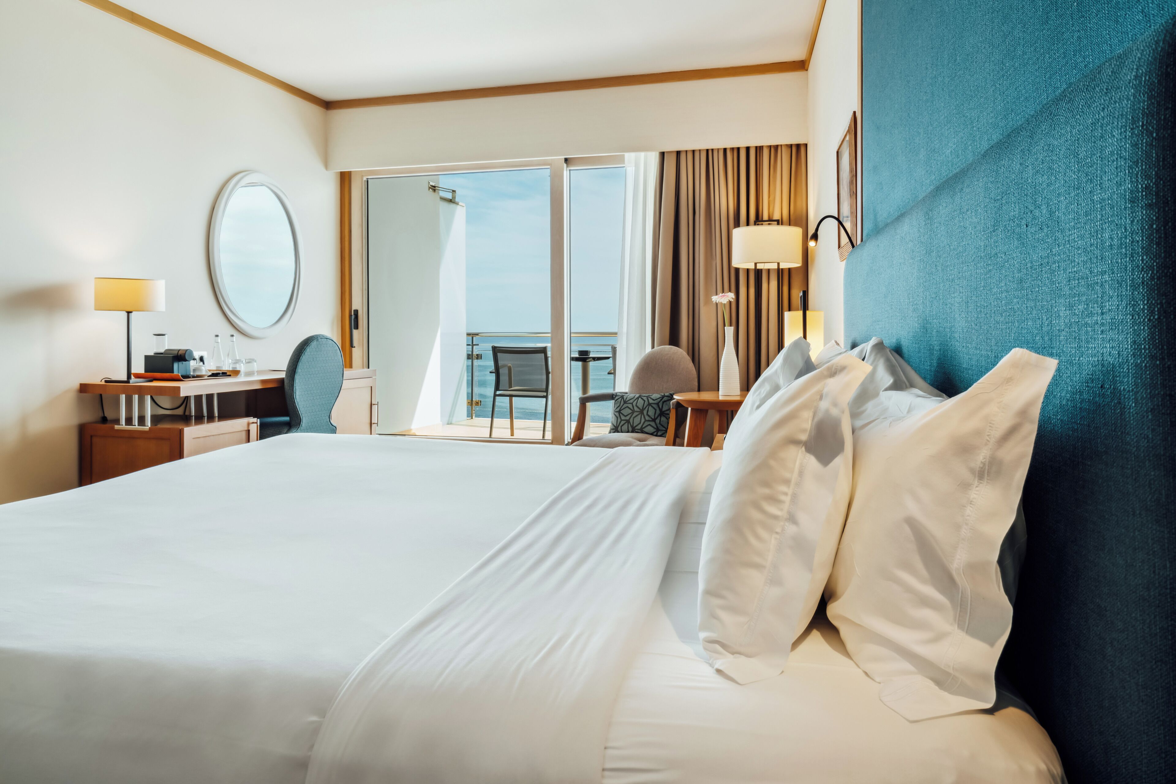 A hotel room with a large bed, a desk with a chair, a round mirror, and a view of the sea through a sliding door.