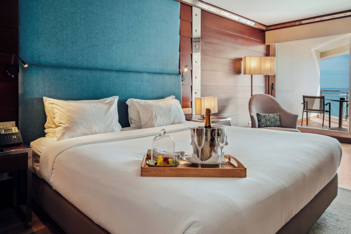 Ein Hotelzimmer mit einem großen Bett, einem Nachttisch mit Telefon, einer Ablage mit Obst und Champagnergläsern sowie Blick auf das Meer.