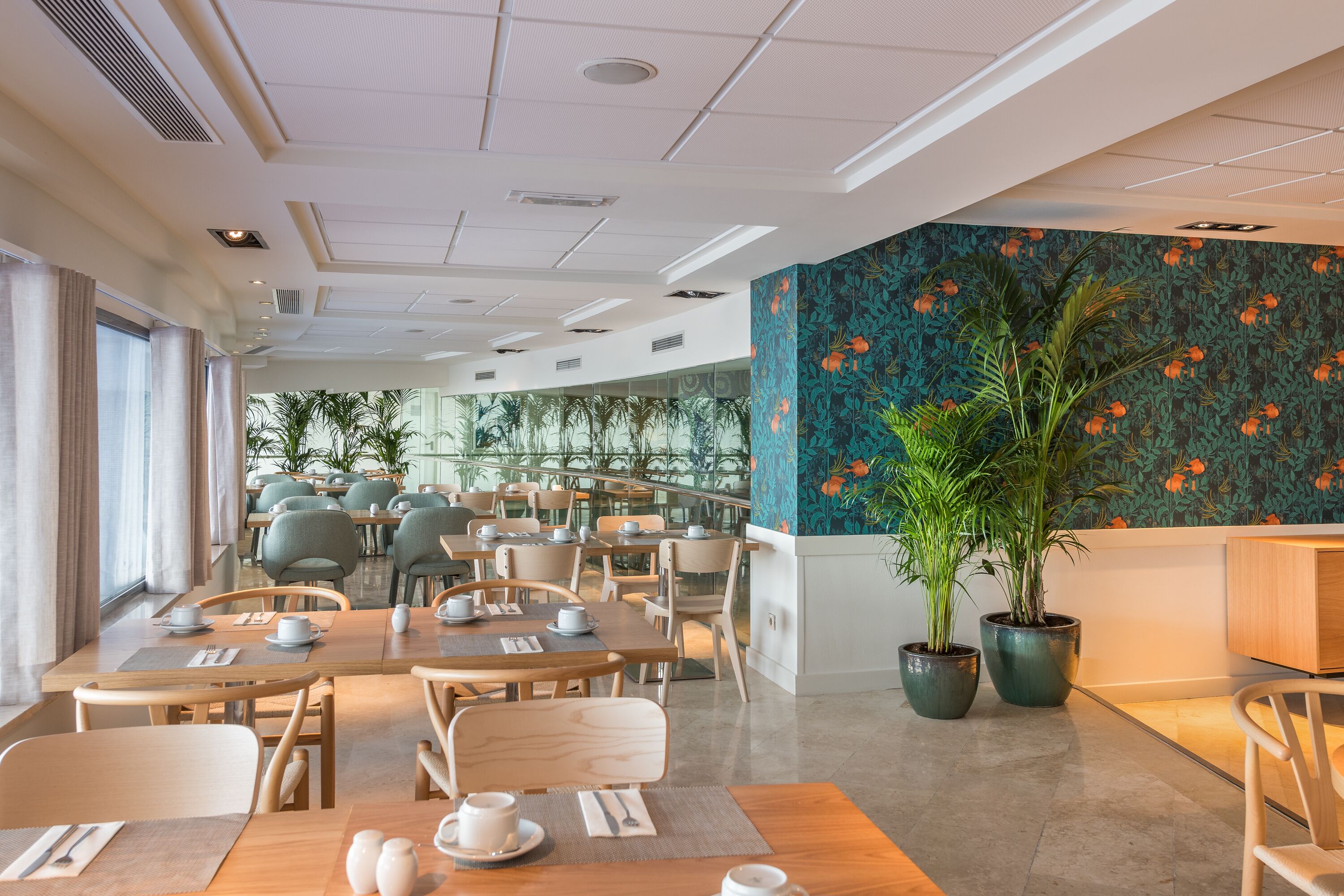 A modern dining area with tables set for service, a large potted plant, and a view of the outdoors through large windows.