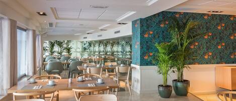 A modern dining area with tables set for service, a large potted plant, and a view of the outdoors through large windows.