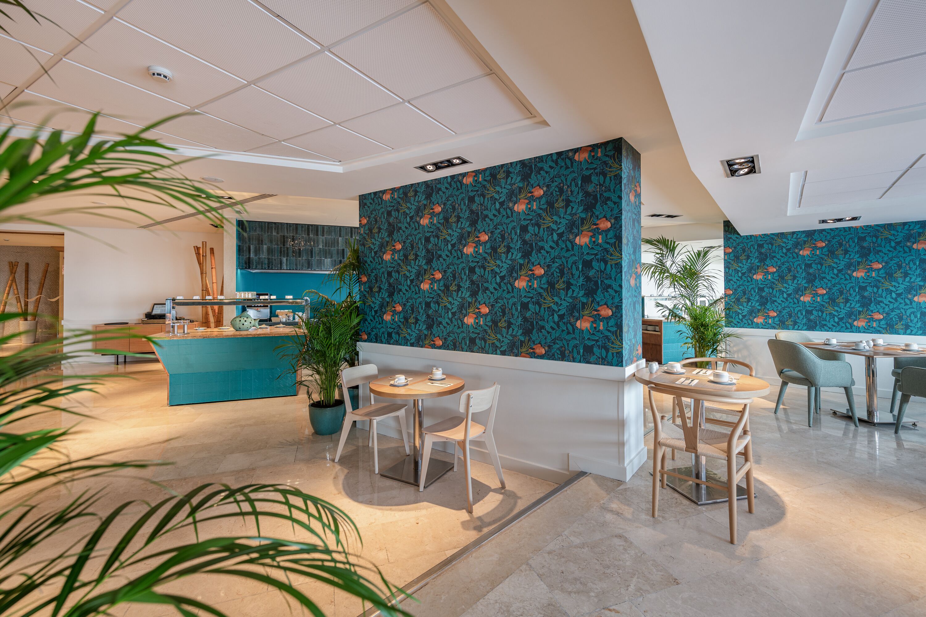 Un hall d’hôtel moderne avec un décor floral aux tons bleus et verts, un comptoir d’accueil et plusieurs espaces de restauration dotés de tables et de chaises rondes.