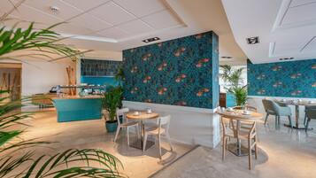 Un moderno vestíbulo de hotel con un diseño floral azul y aguamarina en la pared, un mostrador de recepción y varias áreas de comedor con mesas y sillas redondas.