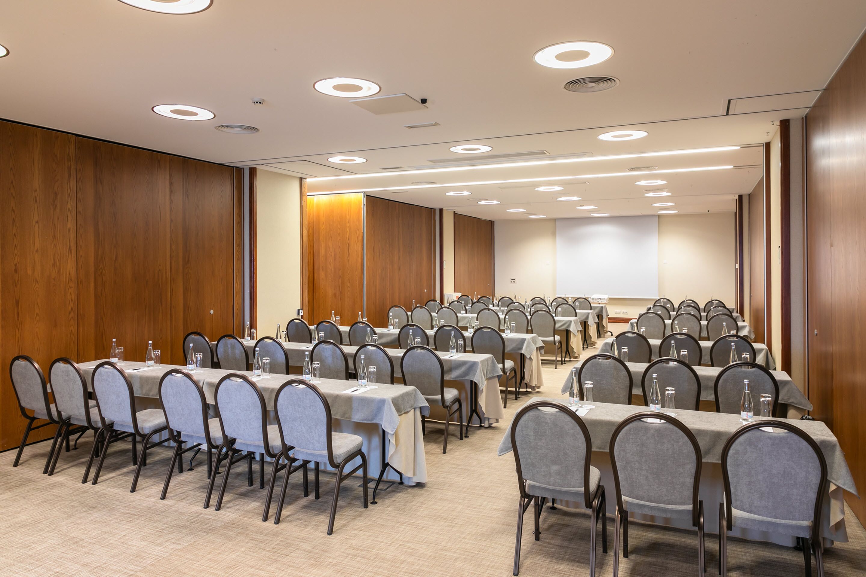 Une salle de conférence dotée de plusieurs tables et de chaises, équipée d’un éclairage au plafond et d’un écran de projection.