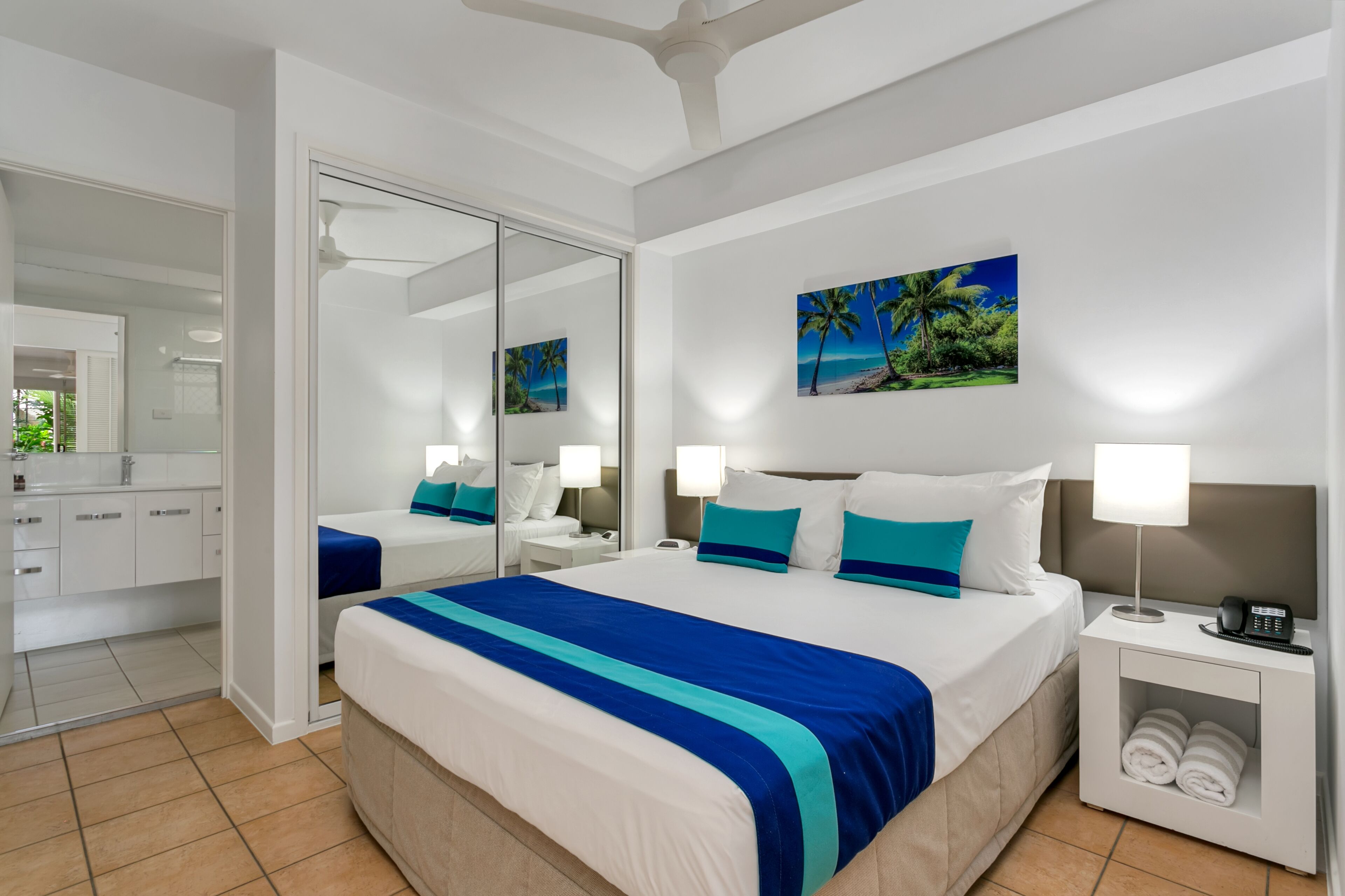 Habitación de hotel con una cama grande, una mesita de noche con lámpara, un espejo y un cuadro con una playa.