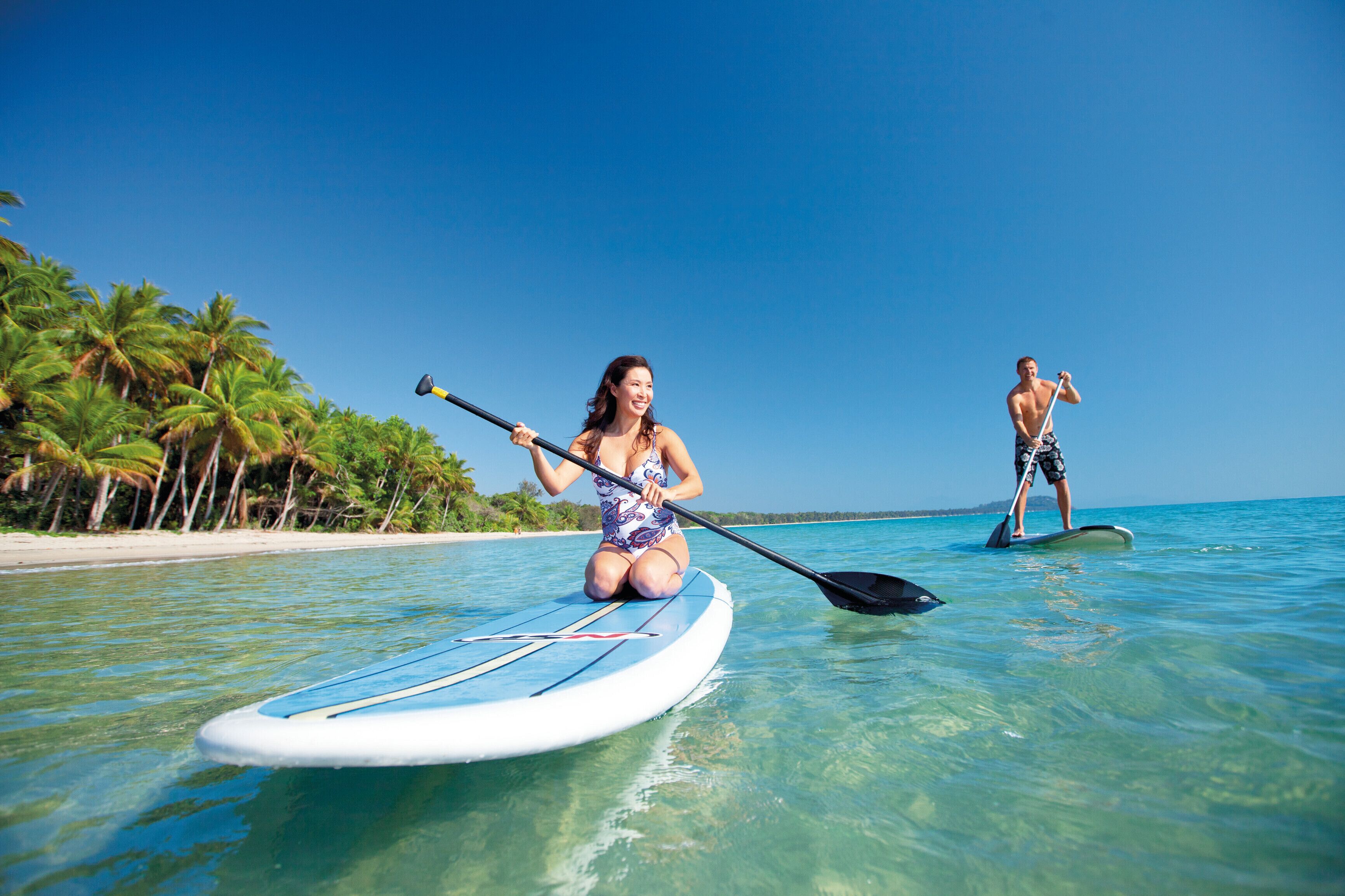 To personer på paddleboards i klart vand med en strand og palmer i baggrunden.