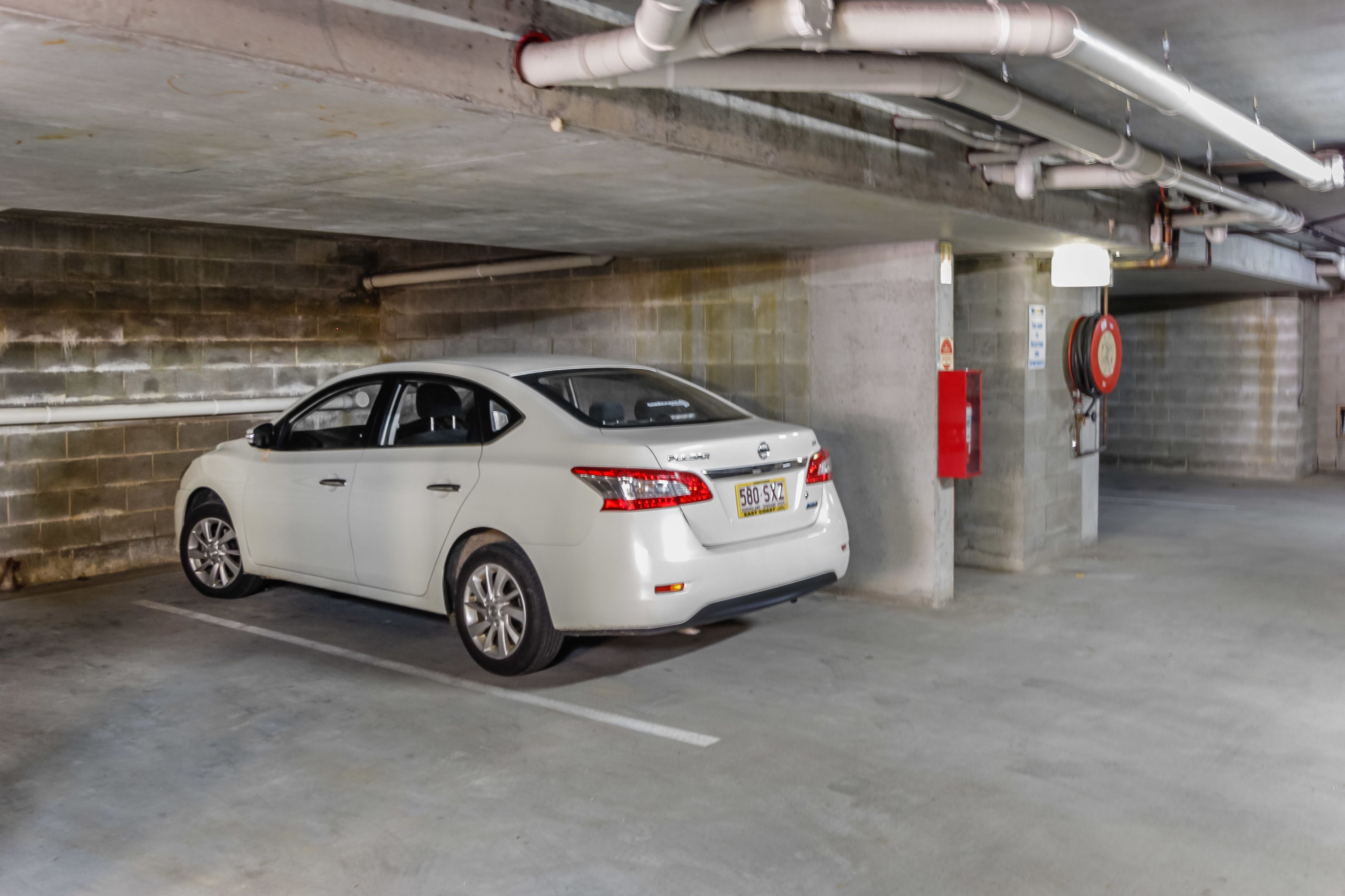 A white car parked in a concrete parking garage with visible pipes and a fire extinguisher.