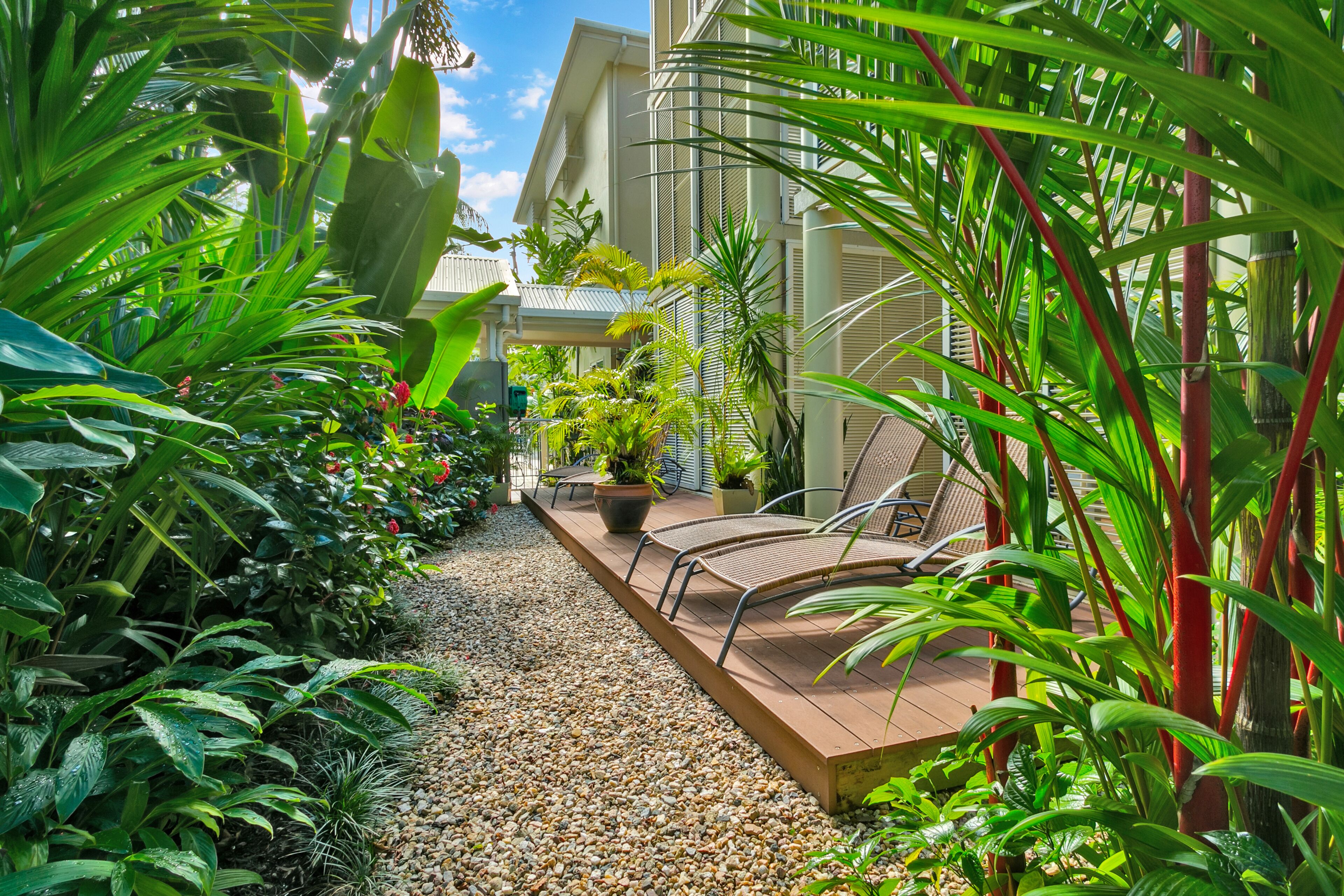 A tropical garden pathway with a wooden deck, potted plants, and a seating area.