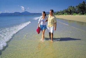 A couple walking on a beach with clear water and a mountain in the background.