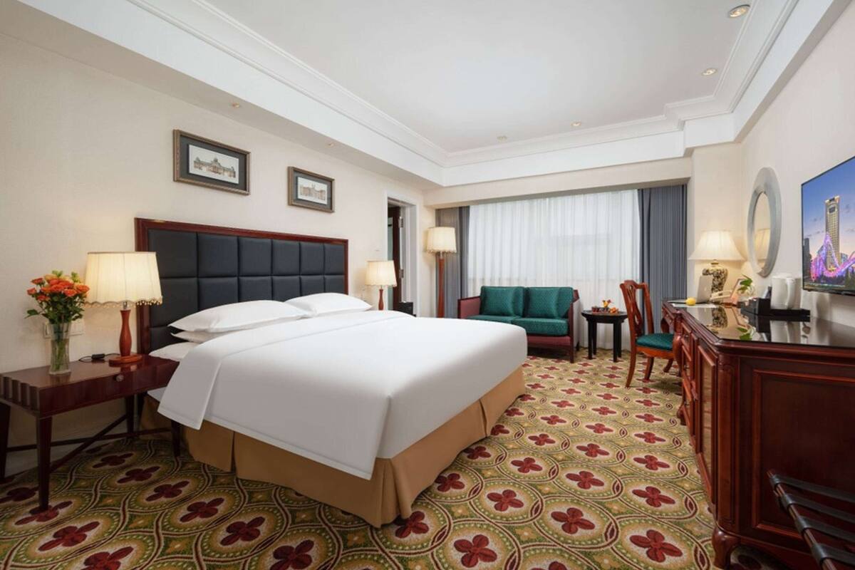A hotel room with a large bed, a desk, a chair, a TV, and a patterned carpet.