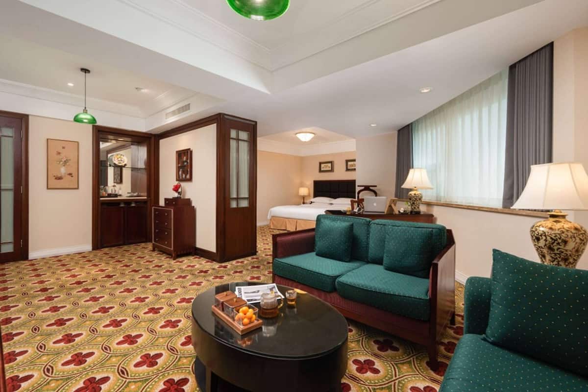 A hotel room with a green sofa, a coffee table with fruit, a bed, a desk, and a large window with curtains.